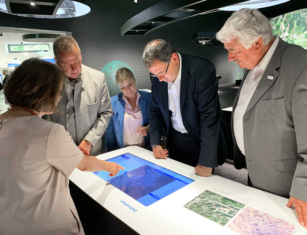 Eröffnung der Sonderausstellung im Bayerischen Landesluftbildzentrum Personen stehen um einen Touch-Bildschirm herum